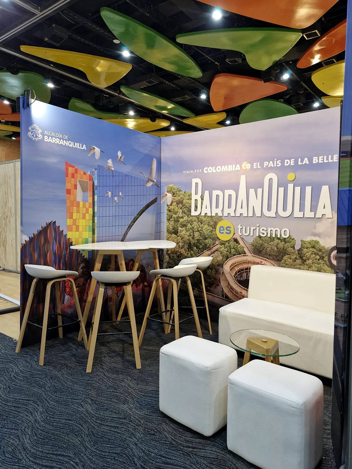 Sala lounge del stand con sofá blanco, dos puffs, mesa de centro en vidrio y mesas altas frente al muro de la Alcaldía de Barranquilla