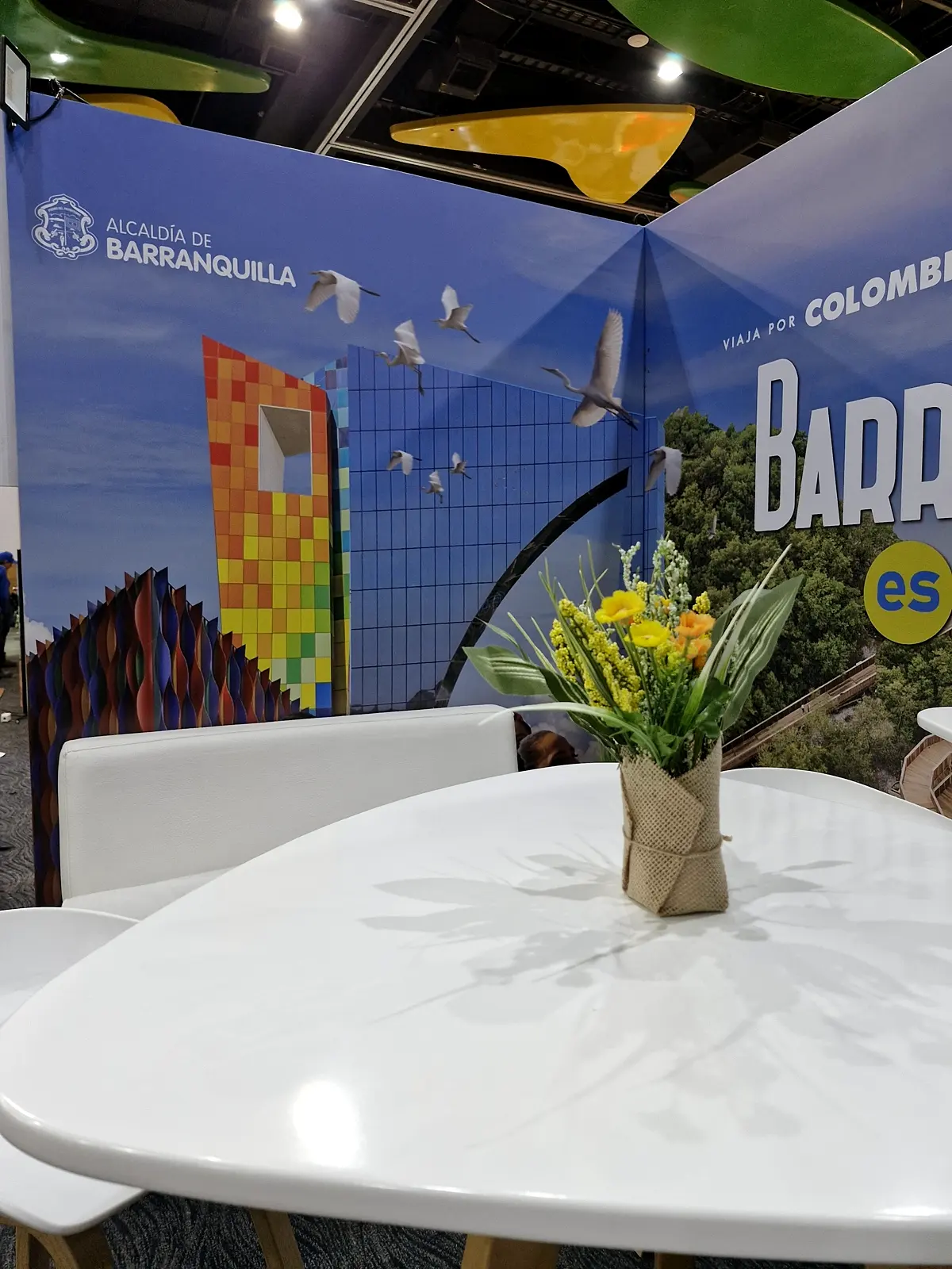 Detalle del muro izquierdo con el logo de la Alcaldía de Barranquilla, edificio de colores y aves, con mesa alta y flores en primer plano