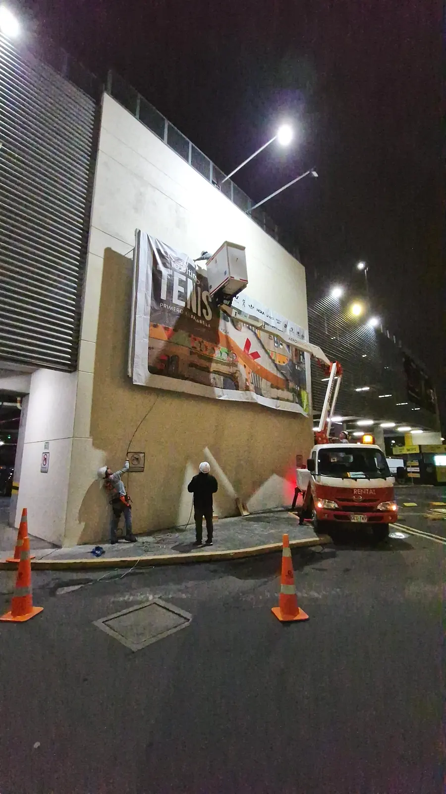 Montaje nocturno de la valla con grúa canasta mientras dos instaladores tensan la lona desde el piso