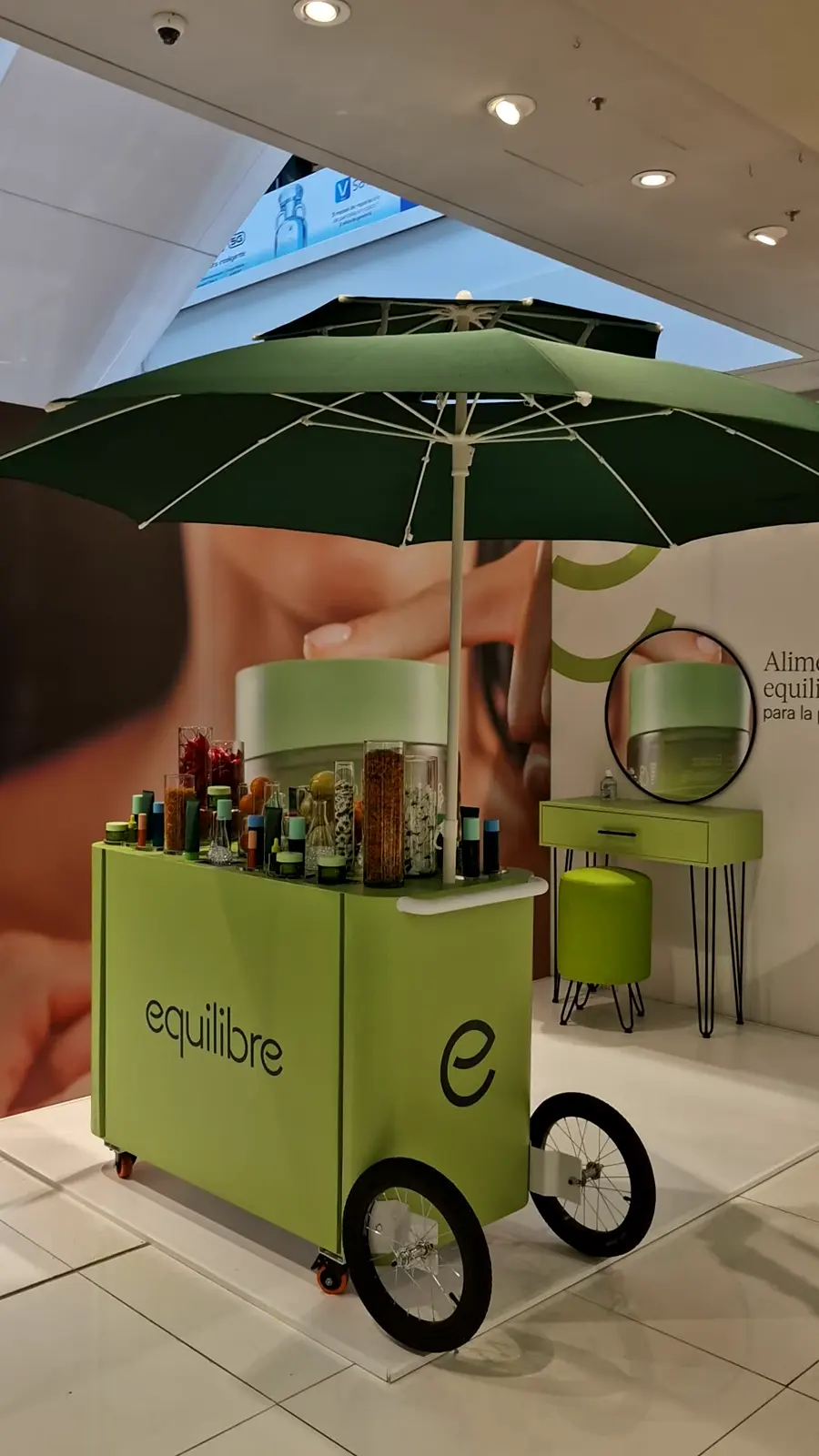Backing en L de Equilibre con carrito verde con sombrilla, tocador con espejo redondo y puff dentro de una tienda Falabella