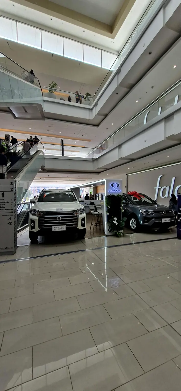 Vista general de la exhibición KGM en el primer piso de Multiplaza: Rexton Sports al frente, tótem SsangYong ambientado y camioneta gris con moño rojo