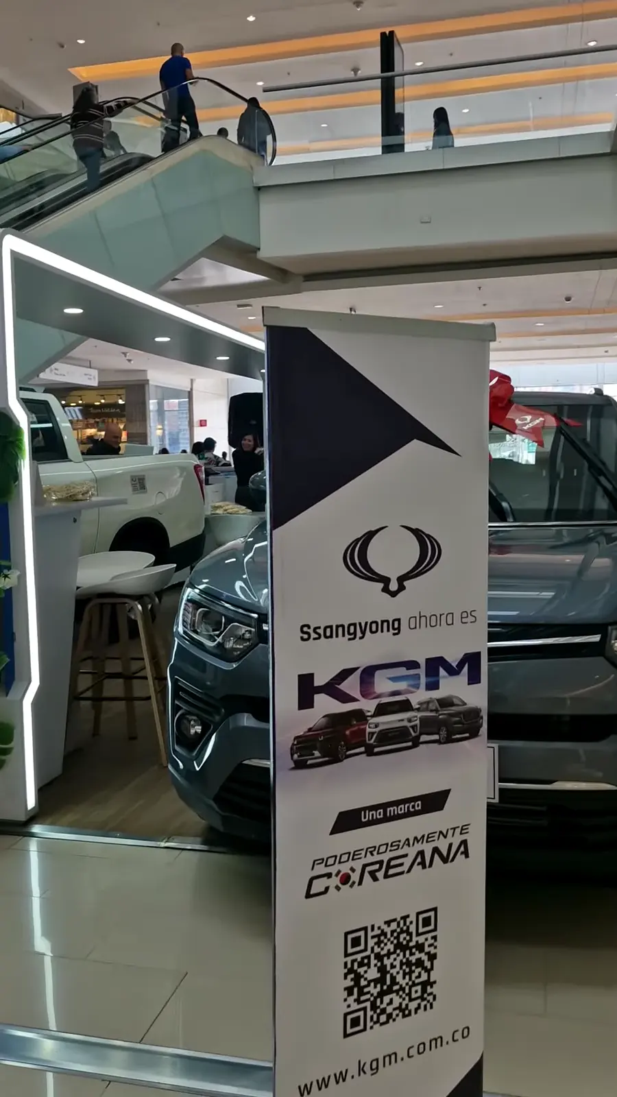 Roll-up con el mensaje «Ssangyong ahora es KGM, Poderosamente coreana» y código QR frente a las camionetas en exhibición
