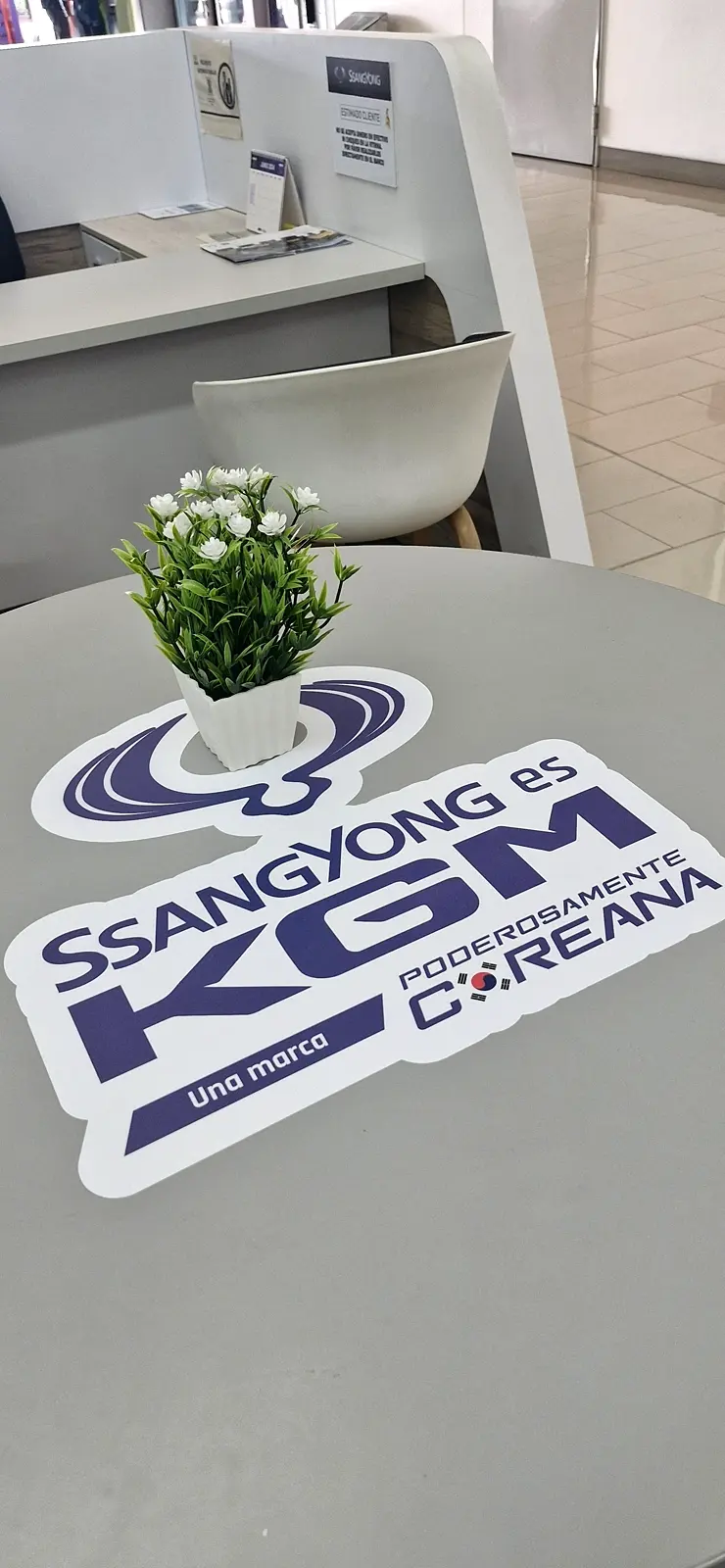 Pieza de mesa troquelada «SsangYong es KGM, Poderosamente coreana» sobre una mesa redonda con una matera de flores blancas