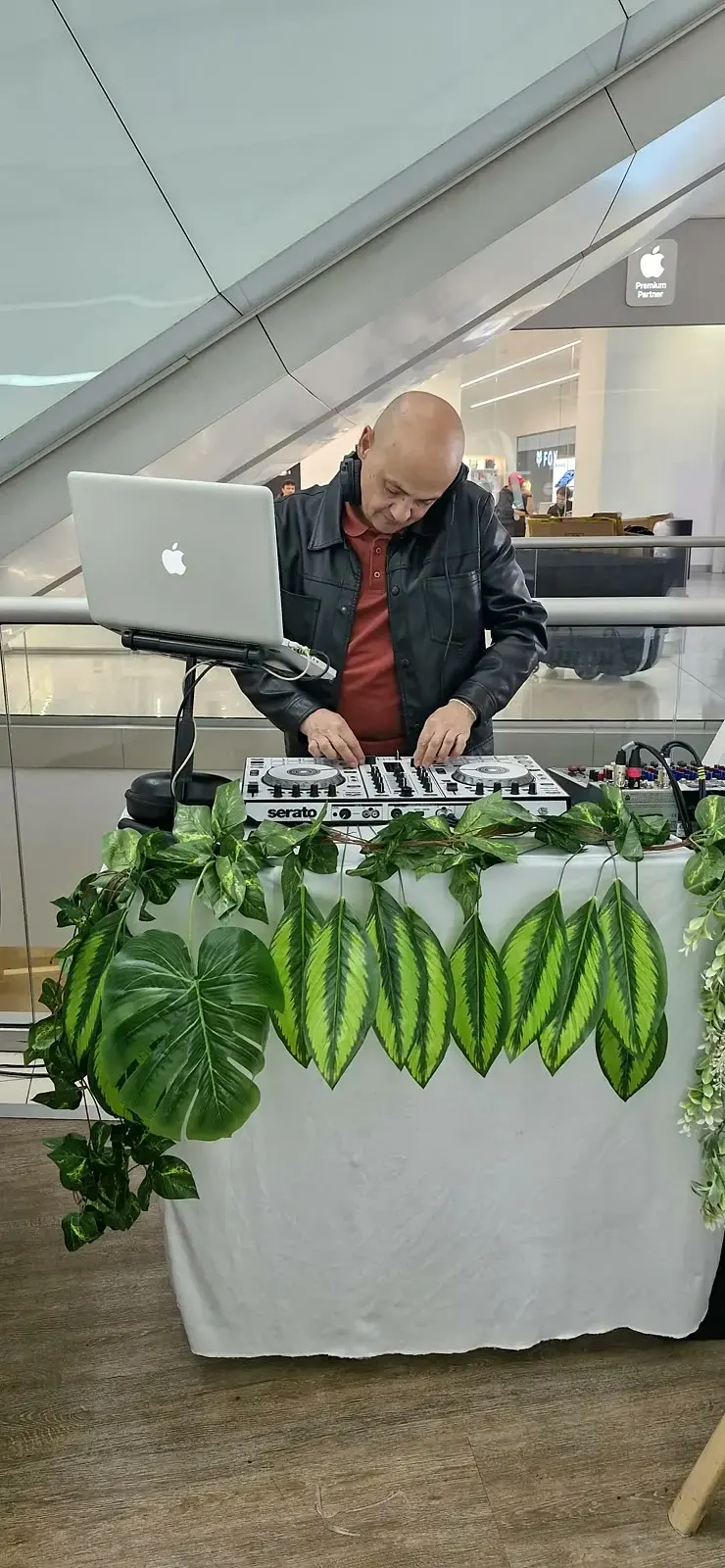 DJ en vivo en una cabina con mantel blanco ambientada con hojas tropicales y enredaderas dentro del centro comercial
