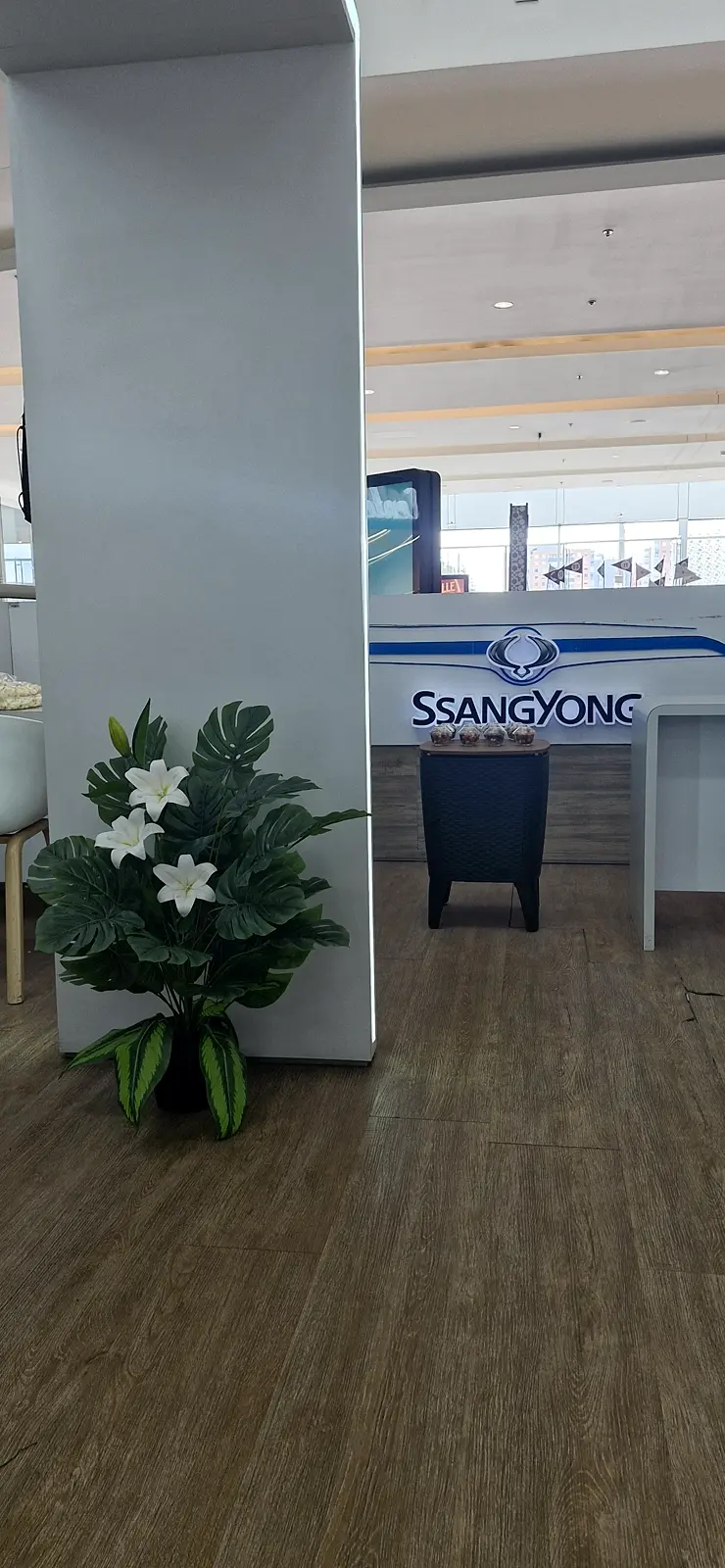 Arreglo de follaje con flores blancas junto a una columna, con el mostrador iluminado de SsangYong al fondo