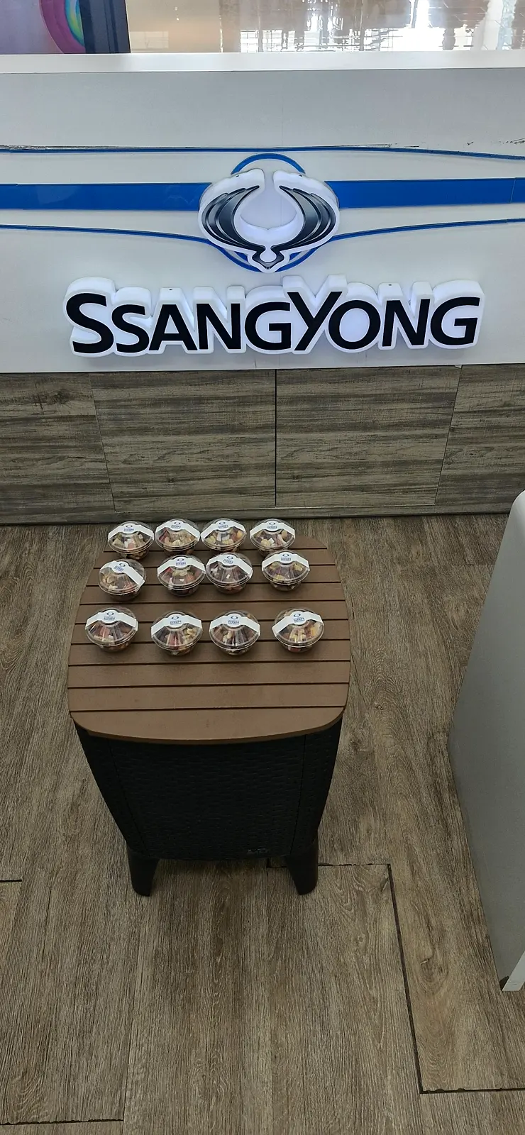 Vasos de pasabocas con etiqueta de marca sobre una mesa auxiliar de madera frente al mostrador iluminado de SsangYong