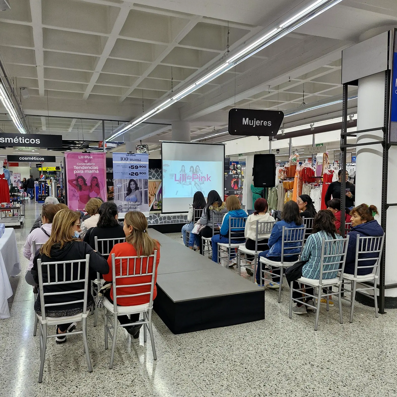 Pasarela recta con público sentado a ambos lados, pendones de Lilipink y pantalla de proyección al fondo, en la sección de mujeres de un almacén de Bogotá