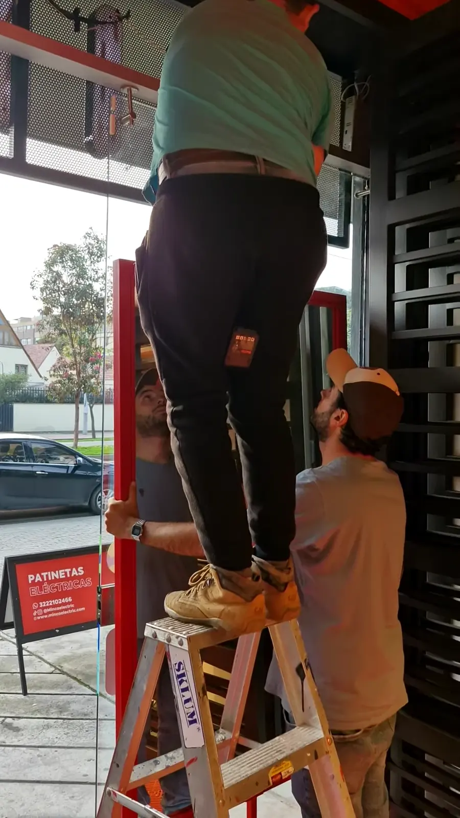Dos instaladores, uno sobre una escalera, montan el tótem en la vitrina de la tienda